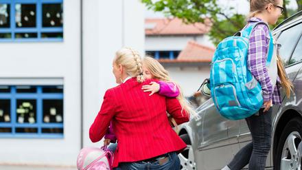 Eine Mutter hockt neben einem Mittelklasse wagen und umarmt ihre Tochter, die eine Schultasche in der Hand hält.