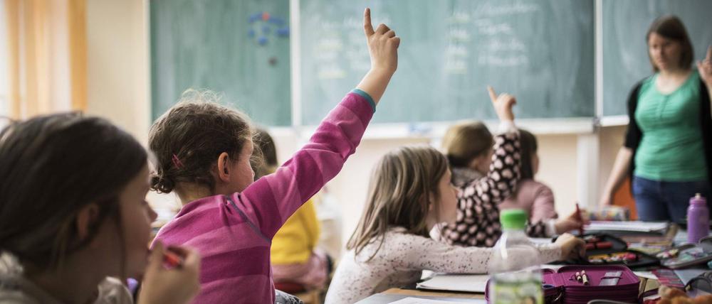 Grundschulkinder sitzen in einem Klassenraum und melden sich, die Lehrerin hebt auch den Finger.