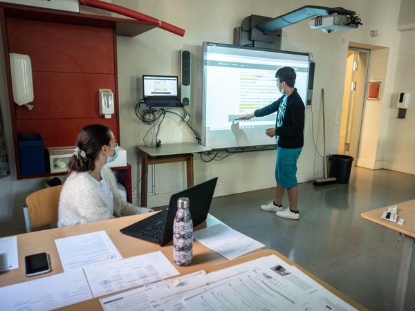 Ein Schüler steht an einem großen Bildschirm vor der Klasse und bearbeitet einen Text, seine Lehrerin sitzt an einem Laptop.