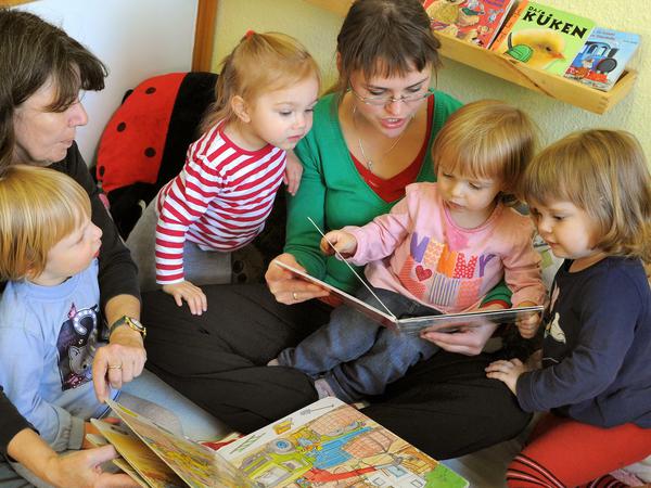 Zwei Kitaerzieherinnen schauen mit vier Kleinkindern in Bilderbücher und sprechen mit ihnen.