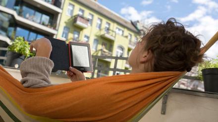 Eine junge Frau liegt auf ihrem Balkon in einer Hängematte und liest ein E-Book.