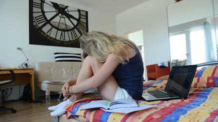  Eine Studentin sitzt auf ihrem Bett in ihrer Wohnung  und legt den Kopf auf ihre Knie. Hinter ihr steht ein aufgeklappter Laptop.