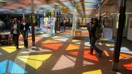 Ein farbiges Glasdach wirft vielfarbige Reflexionen auf den Fußboden einer Eingangshalle, durch die Menschen gehen.