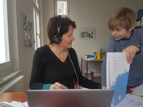 Martina Mörth sitzt mit Headset am Laptop, ihr Sohn zeigt ihr seine Arbeitsblätter.
