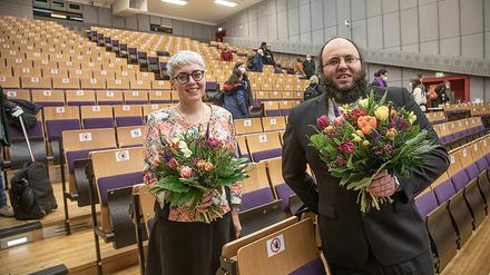Eine Frau und ein Mann stehen mit Blumensträußen in der Hand in einem Hörsaal.