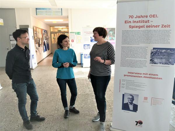 Ein Mann und zwei Frauen stehen an einer Tafel zur Geschichte des Berliner Osteuropa-Instituts und unterhalten sich.