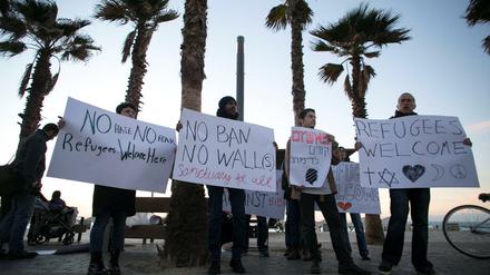 Unter Palmen stehen mehrere Menschen die Plakate mit Aufschriften wie "Refugees Welcome" halten.