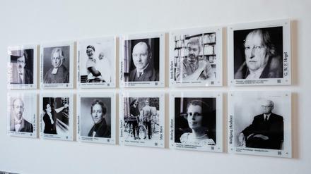 Zwölf Schwarz-Weiß-Porträtfotos historischer Persönlichkeiten sind in einer Zweierreihe an einer Wand angebracht.