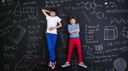 Ein Junge und ein Mädchen liegen lachend auf einer schwarzen Tafel, die mit mathematischen Formeln und Formen vollgeschrieben ist.