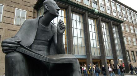 Eine Skulptur vor einem Universitätsgebäude stellt einen sinnierenden Mann mit einem aufgeschlagenen Buch auf den Knien dar.