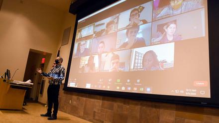 Ein Professor steht in einem Hörsaal, hinter ihm sind auf einer Leinwand Studierende zu sehen, die seine Vorlesung online verfolgen.