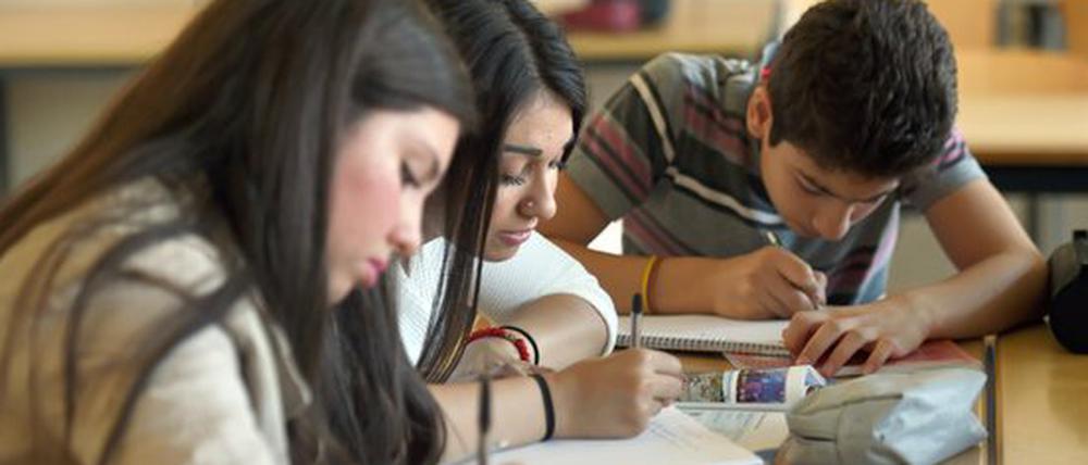 Jugendliche, zwei Schülerinnen und ein Schüler, beugen sich in einem Klassenraum über ihre Kollegblöcke und schreiben.