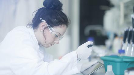 Eine junge Frau arbeitet in einem Forschungslabor, sie gleicht Ergebnisse auf dem Display eines Tablets ab.